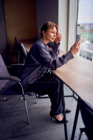                beautiful middle age business woman wearing gray shirt, wide leg pants and black stilettos fix her hair looking into phone camera in modern workspace                                               