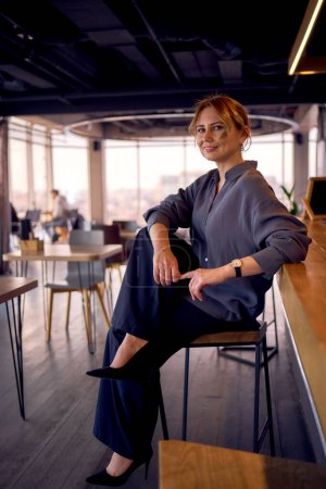 an  attractive middle age woman in a bar with a neutral design, wearing wide leg pants and a silk blouse                       
