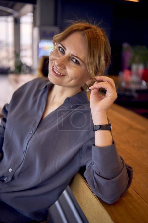 una atractiva mujer de mediana edad en un bar con un diseño neutral, con pantalones de pierna ancha y una blusa de seda                       