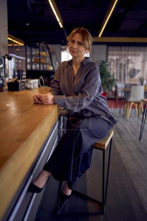  mujer de mediana edad cansada en un bar con un diseño neutral, con pantalones anchos y una blusa de seda                              