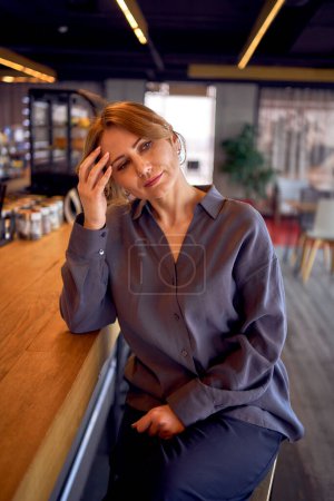  femme d'âge moyen fatiguée dans un bar au design neutre, portant un pantalon large et un chemisier en soie                              