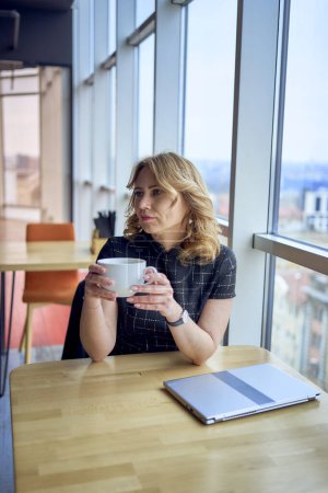    eine atemberaubende reife Frau an einem Tisch in einem Café, das Kaffee in der Nähe von Panoramafenstern mit Blick auf die Stadt trinkt                            