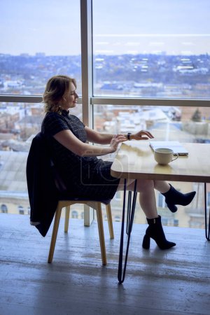    eine atemberaubende reife Frau an einem Tisch in einem Café, das Kaffee in der Nähe von Panoramafenstern mit Blick auf die Stadt trinkt                            