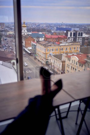                     Person sitting in front of window with high heels, black pants and leg crossed. Cityscape outside. Modern interior with neutral tones.           