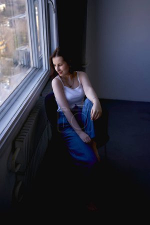 a young teenage girl fighting brain cancer in a studio photo shoot sitting on a chair by the window                      
