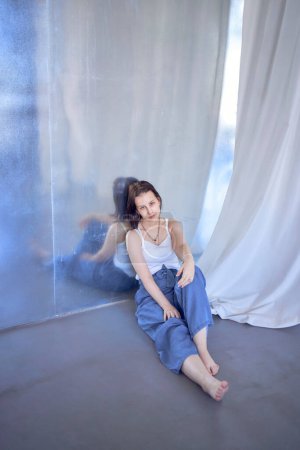 una joven adolescente luchando contra el cáncer de cerebro en la sesión de fotos en el estudio sentado en el suelo, apoyado contra la pared de metal, reflexión