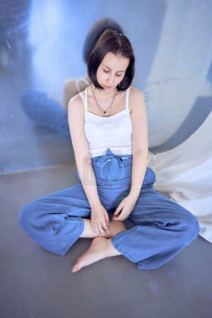 una joven adolescente luchando contra el cáncer de cerebro en la sesión de fotos en el estudio sentado en el suelo, apoyado contra la pared de metal, reflexión