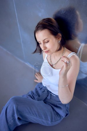 ein junges Teenager-Mädchen, das beim Fotoshooting im Studio gegen Gehirnkrebs kämpft, sitzt auf dem Boden, lehnt sich an eine Metallwand, Spiegelung