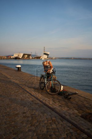 Téléchargez les photos : Burgas, Bulgarie - 17 juin 2023 : un homme répare un vélo après avoir pêché dans le port de Burgas au coucher du soleil - en image libre de droit