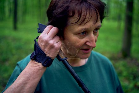  an elderly woman engaged in Nordic walking with sticks in spring forest switches music on headphones                        