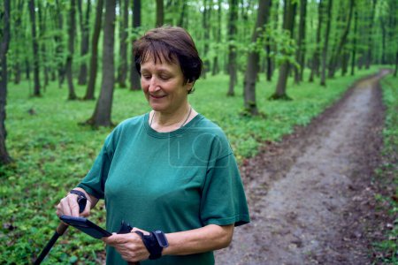  an elderly woman doing Nordic walking with sticks in spring forest switches podcast on phone             