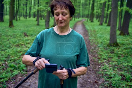 Téléchargez les photos : Une femme âgée faisant de la marche nordique avec des bâtons dans la forêt de printemps commute podcast sur le téléphone - en image libre de droit