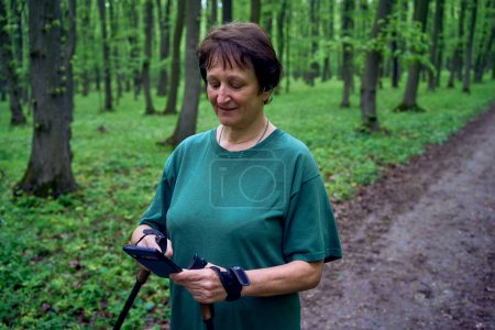  una anciana haciendo marcha nórdica con palos en el bosque de primavera cambia podcast en el teléfono             
