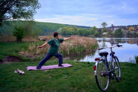 Téléchargez les photos : Une vieille femme pratique le yoga le matin sur la rive de la rivière, est venu de la maison à vélo - en image libre de droit