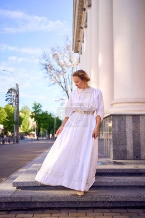 una elegante mujer de mediana edad en vestido vintage blanco cerca del teatro con columnatas antiguas
