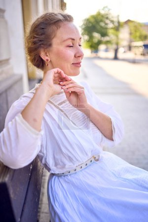 Téléchargez les photos : Une élégante femme d'âge moyen dans une robe vintage blanche assise sur un banc dans la ville du matin - en image libre de droit