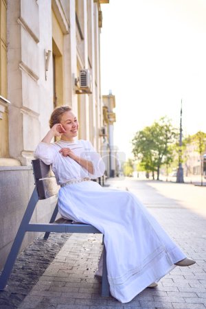 an elegant middle age woman in a white vintage dress sitting on a bench in the morning city
