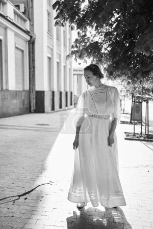 eine elegante Frau mittleren Alters in einem weißen Vintage-Kleid vor dem Hintergrund historischer Gebäude im Morgenlicht