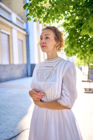 una elegante mujer de mediana edad en un vestido vintage blanco sobre el fondo de edificios históricos en la luz de la mañana