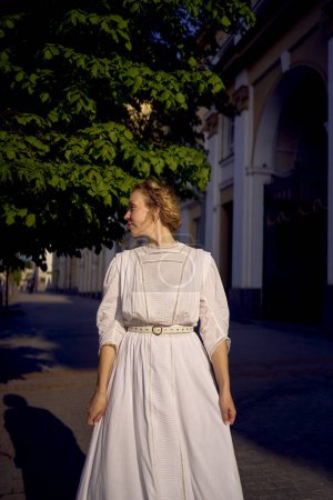 Téléchargez les photos : Une élégante femme d'âge moyen dans une robe vintage blanche sur fond de bâtiments historiques dans la lumière du matin - en image libre de droit