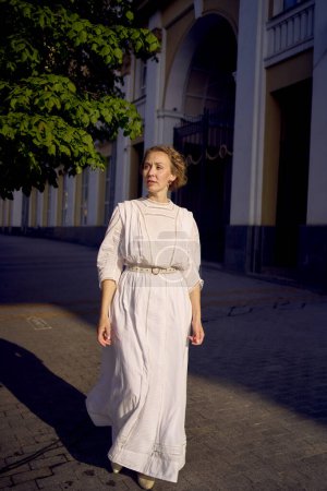 Téléchargez les photos : Une élégante femme d'âge moyen dans une robe vintage blanche sur fond de bâtiments historiques dans la lumière du matin - en image libre de droit