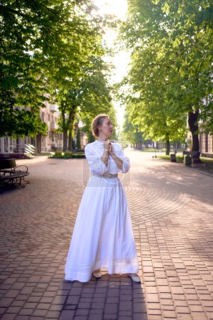 Téléchargez les photos : Une femme chic d'âge moyen dans une robe vintage blanche dans une ruelle ensoleillée - en image libre de droit