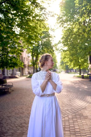 una mujer elegante de mediana edad en un vestido vintage blanco en un callejón iluminado por el sol                      
