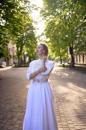 una mujer elegante de mediana edad en un vestido vintage blanco en un callejón iluminado por el sol                      