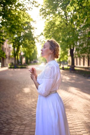 une femme chic d'âge moyen dans une robe vintage blanche dans une ruelle ensoleillée                      