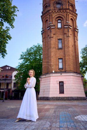 una joven elegante en un vestido vintage blanco en la plaza cerca de la histórica torre de agua en Vinnytsia                  