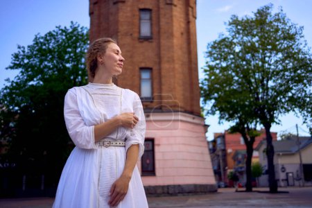 une jeune femme chic dans une robe vintage blanche sur la place près du château d'eau historique de Vinnytsia                  