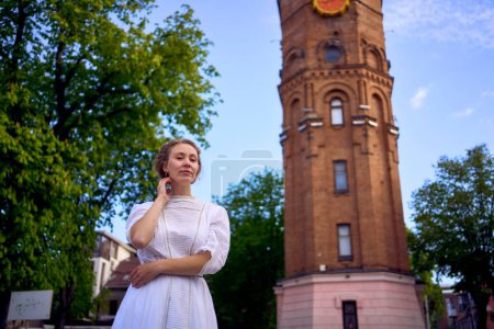 una joven elegante en un vestido vintage blanco en la plaza cerca de la histórica torre de agua en Vinnytsia                  