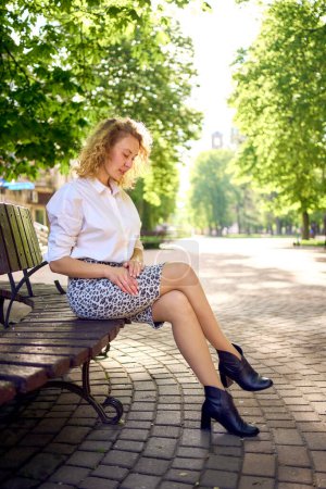 Photo for Beautiful middle age woman in 70s, 80s style clothes on a bench in a sunlit avenue - Royalty Free Image