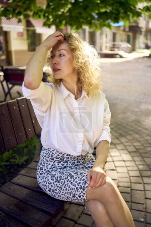 belle femme d'âge moyen dans les années 70, vêtements de style années 80 sur un banc dans une avenue ensoleillée