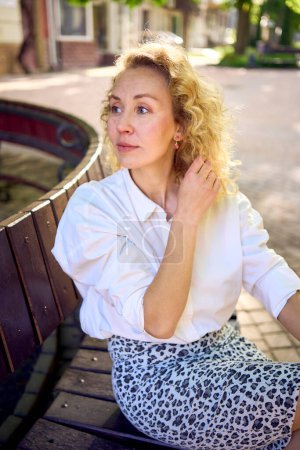 Photo for Beautiful middle age woman in 70s, 80s style clothes on a bench in a sunlit avenue - Royalty Free Image