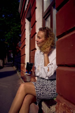 Photo for A beautiful middle age woman in 70s, 80s style clothes with a coffee stain on her shirt drinks coffee sitting on the windowsill - Royalty Free Image