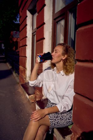 Photo for A beautiful middle age woman in 70s, 80s style clothes with a coffee stain on her shirt drinks coffee sitting on the windowsill - Royalty Free Image