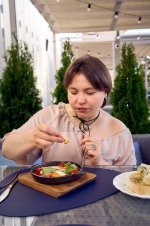 eine mittelgroße Frau im pfirsichflauschigen Kleid, die Shakshouka in einem modernen Restaurant isst