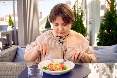 eine mittelgroße Frau im Pfirsich-Fuzz-Kleid isst Quark-Pfannkuchen mit Lachs und Spinat in einem modernen Restaurant                            