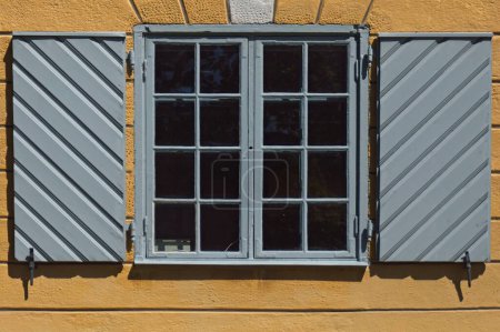 Fenêtre encadrée grise avec couvertures ouvertes en bois sur un vieux bâtiment en pierre jaune.