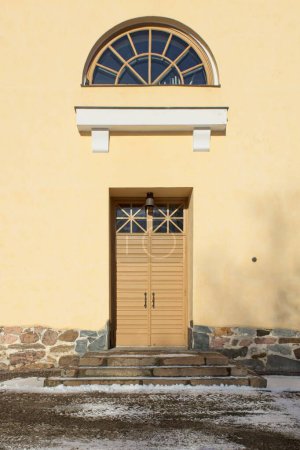 Fenêtre encadrée peinte en brun clair et portes doubles en bois sur un bâtiment en pierre peinte en jaune.