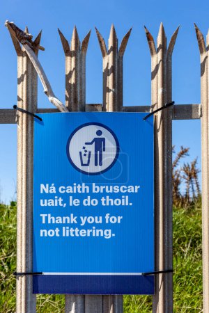 Blue, bring your litter home sign on a metal fence. There are bins, trees and grass behind the fence with a blue sky and no clouds
