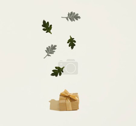 Eine Geschenkschachtel mit grünen Blättern, die darüber auf beigem Hintergrund schweben. Kopierraum. Minimales Konzept von Einkaufen, Urlaubsvorbereitung.