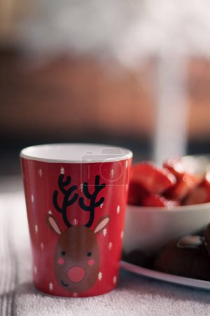 A festive Christmas child's reindeer cup with a plate of strawberries in the back.