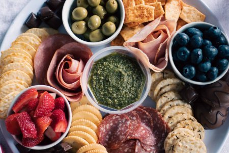 An artisanal, antipasto, fresh fruit, meat, and cracker grazing board finger food, with strawberries, blueberries, olives, ham, salami, chocolate and crackers.