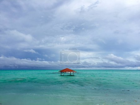 Stunning image of an abandoned red-roofed gazebo flooded by seawater due to abrasion against a backdrop of turquoise sea and cloudy blue sky.