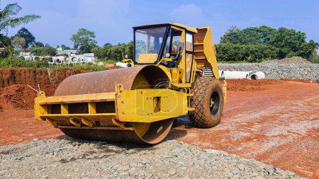 Photo for A yellow roller construction machine working on a road construction project. Plenty of red soil and crushed stone with clear blue sky on the background - Royalty Free Image