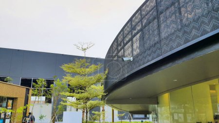 Téléchargez les photos : Jakarta 01 Nov 2023 : Bâtiment café en forme de ruban gris avec façade perforée et vitrée. forme curviligne de torsion unique. iconique. ciel bleu, aménagement paysager luxuriant. - en image libre de droit