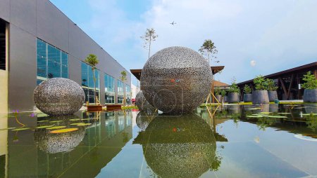 Téléchargez les photos : Jakarta 01 Nov 2023 : Plan frontal d'une sphère métallique flottant sur un étang réfléchissant. nénuphars, plantes tropicales. ballons, ciel couchant. Indonésie Design District centre commercial de bande extérieure - en image libre de droit