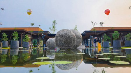 Téléchargez les photos : Jakarta 01 Nov 2023 : Plan frontal d'une sphère métallique flottant sur un étang réfléchissant. nénuphars, plantes tropicales. ballons, ciel couchant. Indonésie Design District centre commercial de bande extérieure - en image libre de droit
